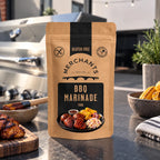 A stand-up pouch of Merchants BBQ Marinade stands on a dark countertop in a kitchen setting, with grilled food, a grill, and bowls of ingredients blurred in the background.
