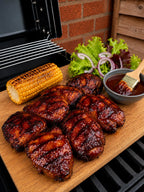 Grilled chicken pieces with a glossy barbecue sauce, served on a wooden cutting board with a grilled corn cob, lettuce, red onion, and a small bowl of sauce with a brush, set on a grill in front of a brick wall.