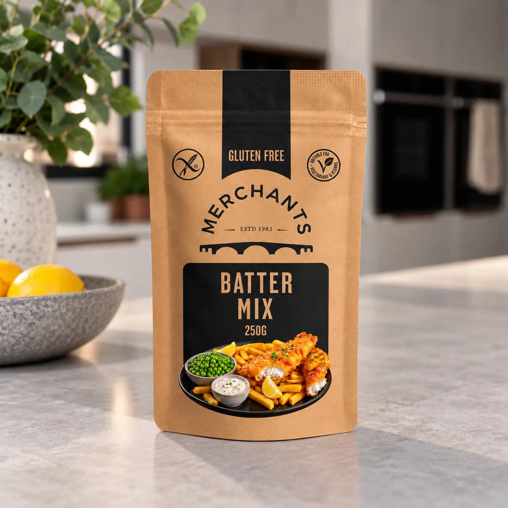 A stand-up pouch of Merchants Batter Mix, 250g, set on a light gray kitchen countertop with a blurred background of a kitchen, a bowl of lemons, and a potted plant.