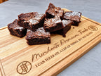 A pile of chocolate brownies dusted with powdered sugar sits on a wooden cutting board engraved with the text 'Merchant's Premium Foods I CAN TOLERATE A LOT OF THINGS, BUT NOT GLUTEN'.