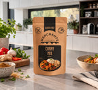 A stand-up pouch of Merchants Curry Mix stands on a kitchen countertop, surrounded by cooking ingredients and a bowl of prepared food in a bright, modern kitchen setting.