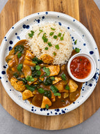 A plated meal featuring a chicken and egg curry with spinach, served over white rice with cilantro garnish, alongside a small side of red chili sauce, all on a speckled white plate over a wooden board.