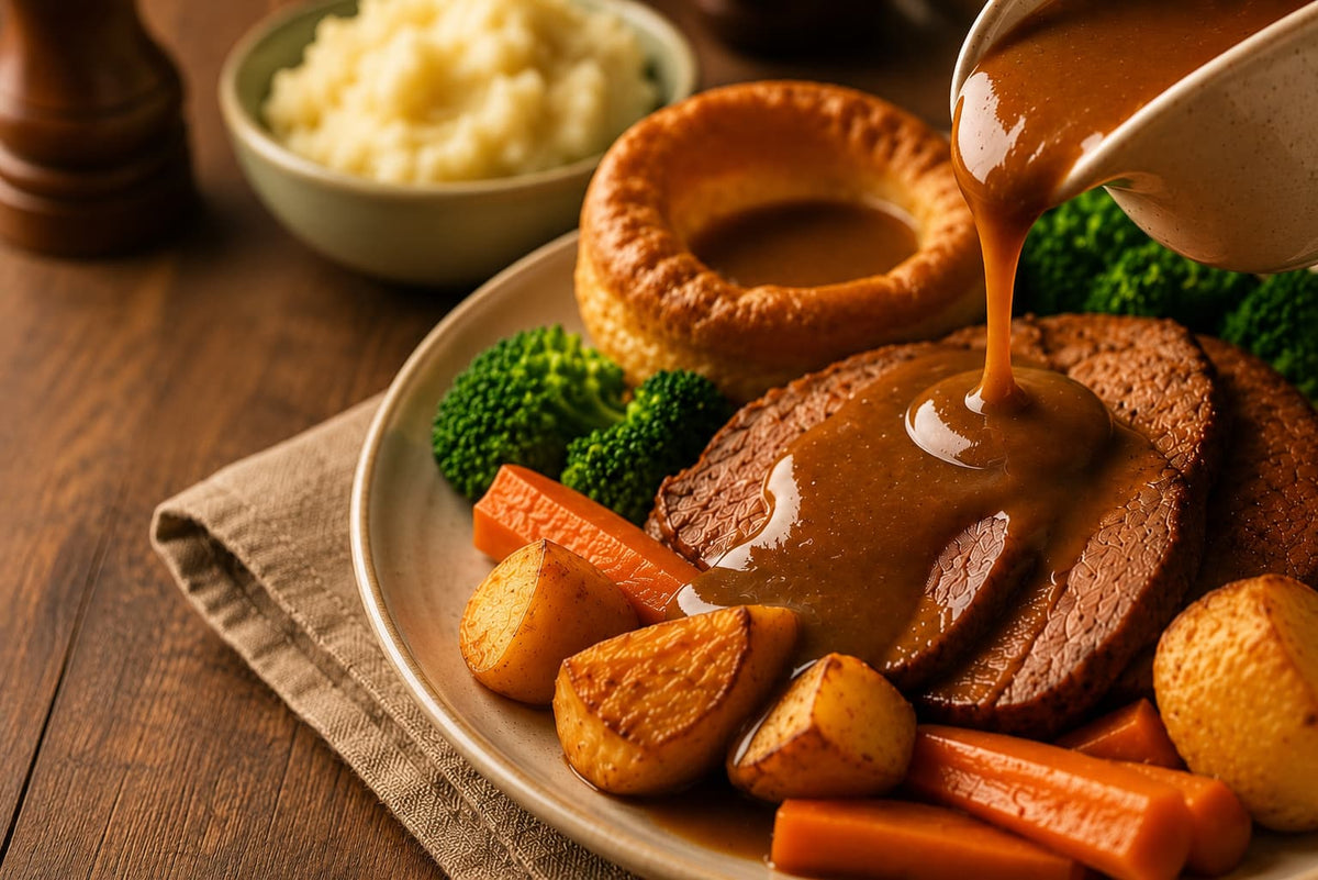 A close-up of a plated meal with sliced roast beef topped with brown gravy, a Yorkshire pudding, broccoli, carrots, roasted potatoes, and mashed potatoes on a wooden table.
