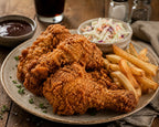 A plate of golden-brown fried chicken pieces served with french fries, coleslaw, and two dipping sauces on a rustic wooden table.