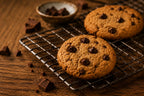 Two chocolate chip cookies cooling on a metal wire rack over a wooden table, with scattered chocolate chunks and a small bowl of chocolate pieces in the background.