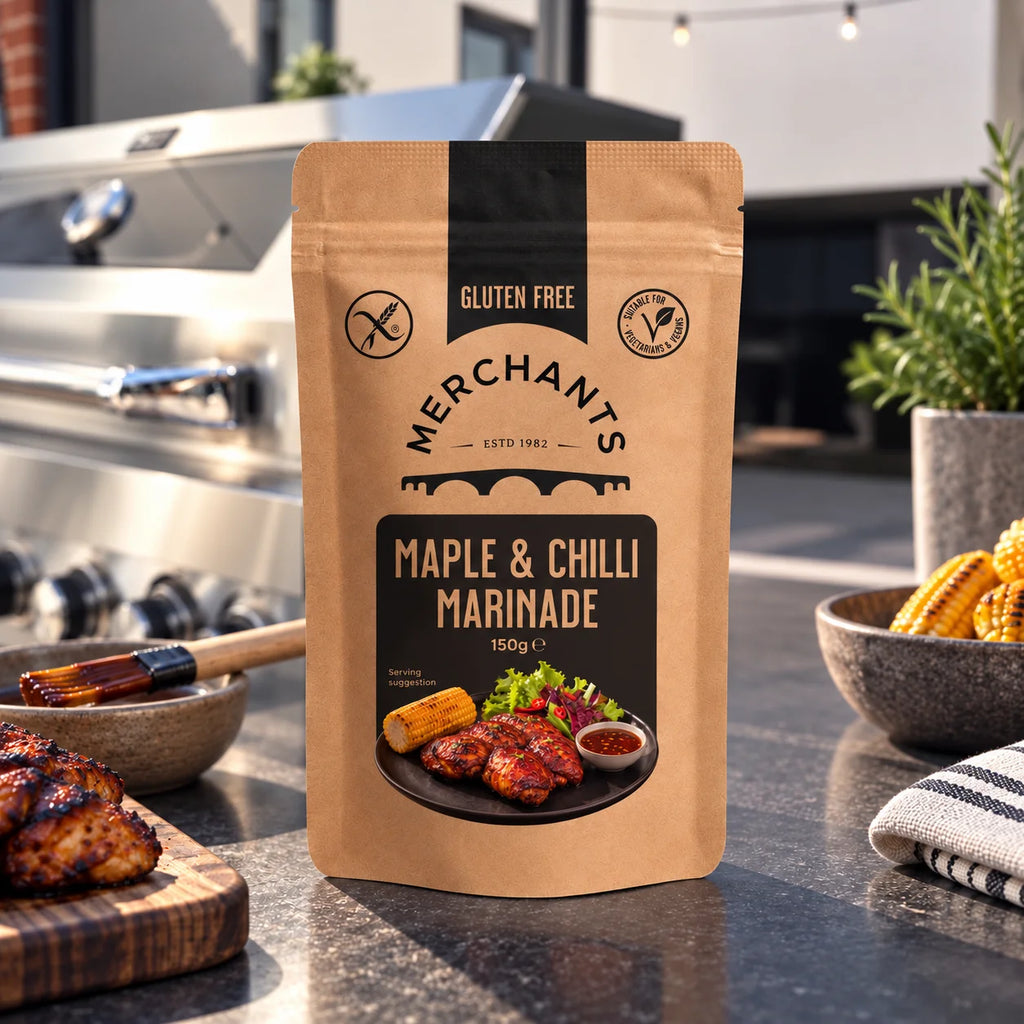 A stand-up pouch of Merchants Maple & Chilli Marinade stands on a dark countertop in a kitchen setting, with grilled food, a grill, and bowls of ingredients blurred in the background.