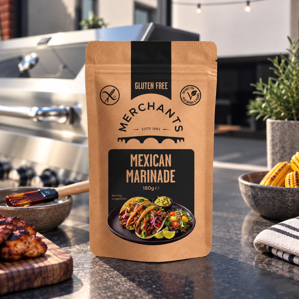 A stand-up pouch of Merchants Gluten Free Mexican Marinade stands on a dark countertop in a kitchen setting, with grilled food, bowls, and a grill visible in the background.