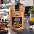 A stand-up pouch of Merchants Gluten Free Mexican Marinade stands on a dark countertop in a kitchen setting, with grilled food, bowls, and a grill visible in the background.