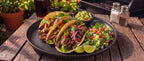 Three soft corn tacos filled with grilled meat and topped with red onion, cilantro, and diced tomatoes, served on a black plate with guacamole, a side salad, lime wedges, and a drink on a rustic wooden table outdoors.