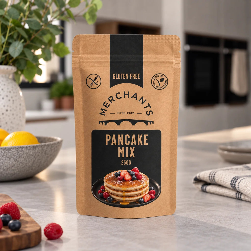A stand-up pouch of Merchants Pancake Mix, 250g, placed on a light gray kitchen countertop with a blurred kitchen background, surrounded by fresh berries, a lemon, and a checkered cloth.