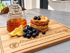 A wooden serving board engraved with a humorous quote and logo, styled with a glass honey pot, lemon slices, blueberries, and a stack of pancakes on a kitchen countertop.
