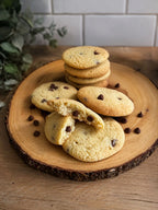 Chocolate chip cookies arranged on a round wood slice platter, with one cookie broken in half to show the interior texture. The cookies are golden brown with dark chocolate chips, set on a wooden table with a white tiled backsplash in the background.