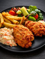 A plated meal featuring two golden-brown breaded chicken pieces, french fries, a potato salad, and a fresh salad with tomatoes, cucumber, and greens on a black plate.